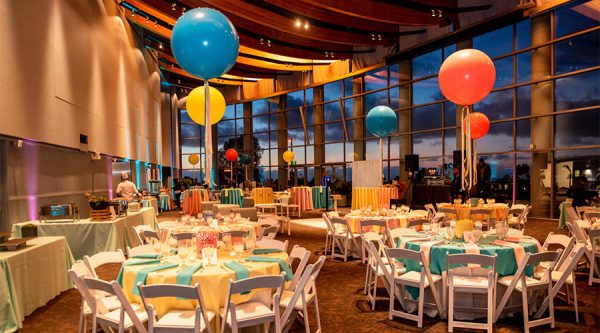 A picture of an elegant banquet with round tables, balloon centerpieces, and a nighttime view of the Pacific Ocean.